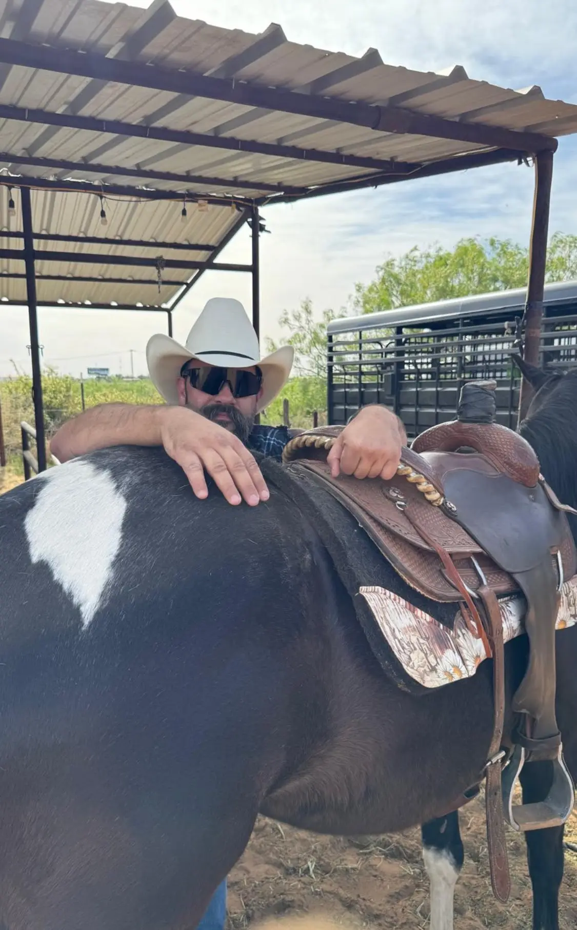 Un cowboy col cappello bianco appoggiato alla sella di un cavallo pezzato